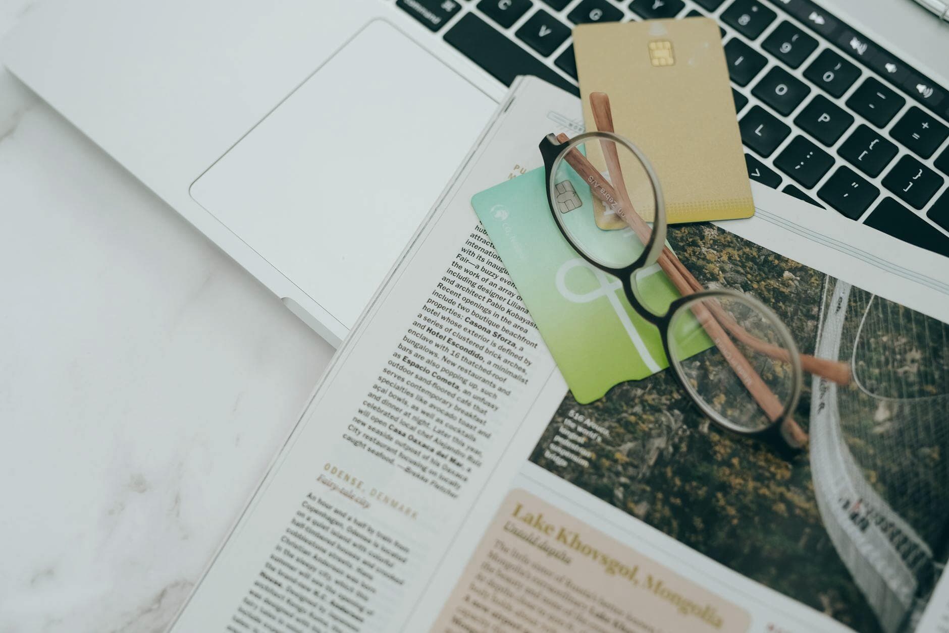 Eyeglasses resting on a magazine beside a laptop and credit cards, indoors.